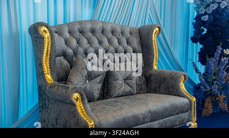 Luxurious vintage velvet sofa featuring tufted upholstery and decorative cushions, set against a backdrop of elegant blue drapes and floral decor, per Stock Photo