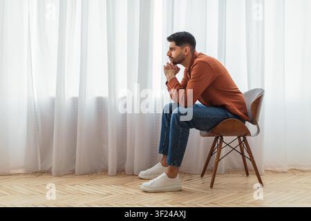 Thoughtful Young Man Sitting On Chair Looking Out Window, Focused On Daydreaming Concept Stock Photo