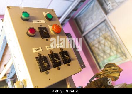 Close-up of an industrial machine control panel featuring various switches, buttons, and emergency stop, set in a factory environment with machinery. Stock Photo