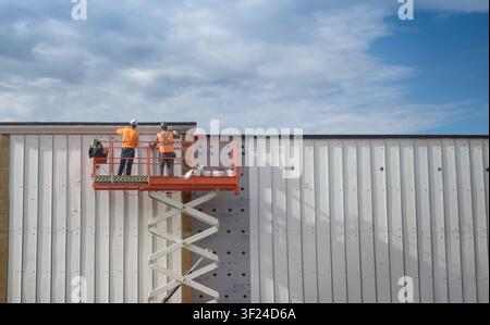 Two construction workers on a scissor lift installing insulated metal wall panels on a commercial building exterior under a dramatic cloudy sky. Indus Stock Photo