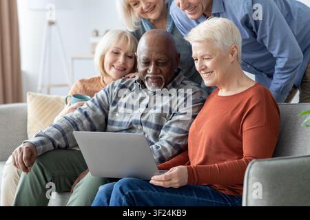Happy Senior Couple Using Laptop Together With Friends In Living Room, Technology Concept Stock Photo