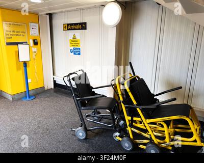 Stacked airport wheelchairs and mobility aids in a Manchester arrivals area beside signage and lift. Stock Photo