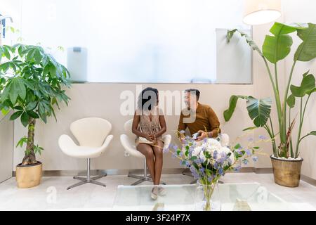 Diverse couple sitting and talking in a modern health clinic waiting area with houseplants Stock Photo