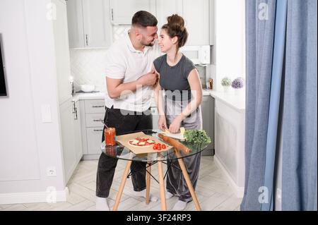 Smiling young man and woman hugging, looking at each other and laughing while preparing homemade pizza at a glass table during a cozy romantic date an Stock Photo