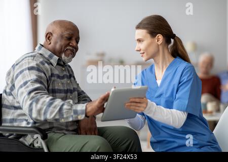 Elderly African American Man In Wheelchair Interacting With Female Nurse Using Tablet, Healthcare Concept Stock Photo