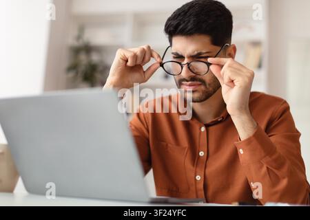 Frustrated Young Man Wearing Glasses Working On Laptop In Bright Office, Focused On Screen Stock Photo