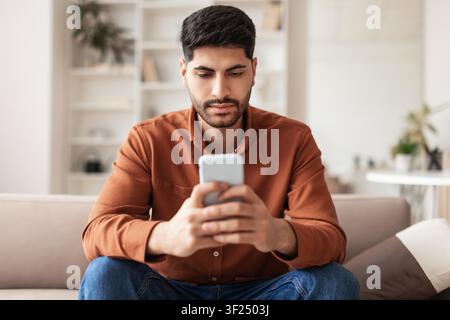 Focused Middle Eastern Man Using Smartphone In Modern Living Room, Technology Concept Stock Photo