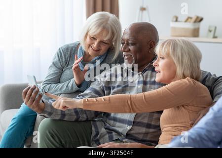 Happy Senior Caucasian Women And Black Man Using Smartphone Together At Home, Family Bonding Concept Stock Photo