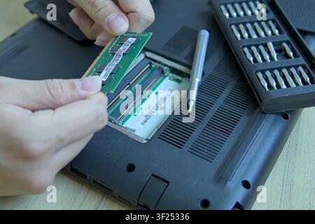 Close-up of a person's hands holding a green DDR memory module, preparing to push it into the laptop's empty motherboard RAM slot. A precision screwdr Stock Photo