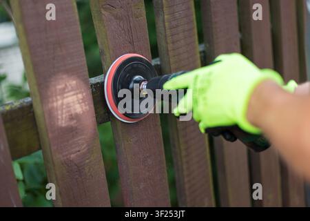 Close-up of worker in bright protective gloves sanding a rustic wooden fence using a cordless drill power tool. Home renovation, DIY, and backyard mai Stock Photo