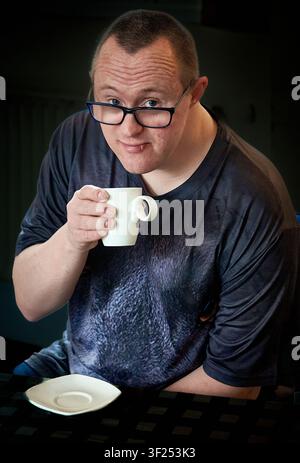 Portrait of a young man with Down syndrome holding a cup of coffee and smiling at the camera, UK Stock Photo