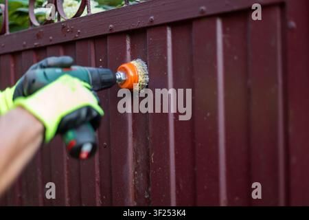 Worker using a cordless drill with a wire brush attachment to clean or strip paint from a painted maroon metal gate. Wearing yellow and black safety g Stock Photo