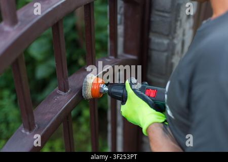 Man in bright safety gloves cleans an old metal gate from rust using an electric cordless drill with a wire brush cup attachment. DIY home renovation, Stock Photo