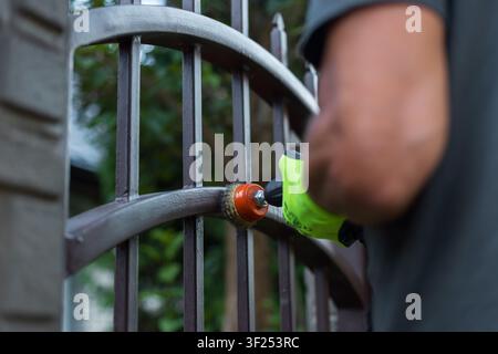 Worker in green safety gloves uses an electric cordless drill with a wire brush attachment to clean and restore an old metal fence. DIY home backyard Stock Photo