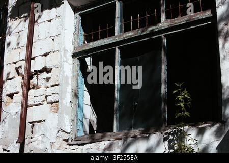 A weathered industrial building with broken windows, cracked masonry, and rusted metal bars, gradually being reclaimed by nature Stock Photo