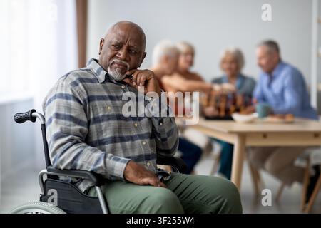 Thoughtful Elderly Black Man In Wheelchair At Social Gathering, Engaging With Friends Stock Photo