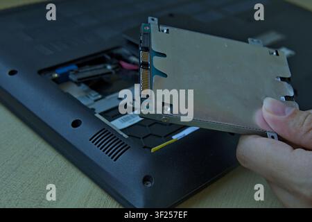 Close-up of a hand carefully removing a 2.5-inch SATA hard disk drive (HDD) in its metal bracket from an open laptop bay. Stock Photo