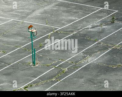 Abandoned basketball court featuring a weathered wooden chair on a green metal pole, worn asphalt surface, overgrown grass, faded white lines, isolate Stock Photo