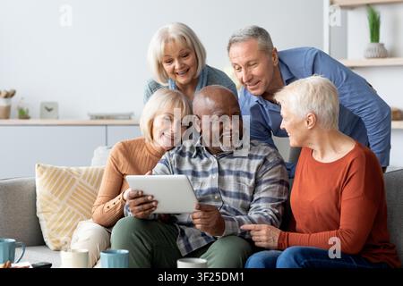 Happy Senior Group Of Friends Smiling And Sharing Digital Tablet At Home, Social Connection Concept Stock Photo