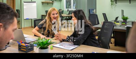 Diverse business woman laughing together while viewing phone screen in coworking office Stock Photo
