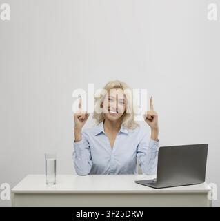 Smiling Young Caucasian Woman Sitting At Desk With Laptop, Water Glass, Pointing Upward, Business Concept Stock Photo