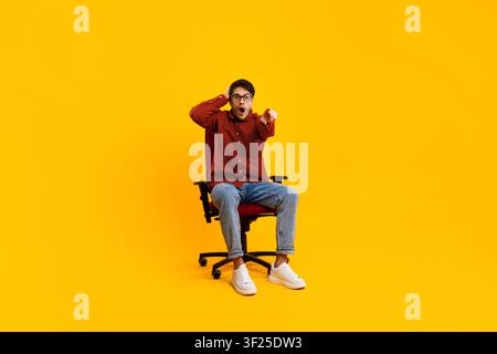 Excited Young Man In Casual Clothes Sitting On Chair And Pointing Finger, Yellow Background Stock Photo
