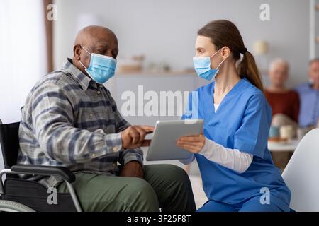 African American Man In Wheelchair Engaging With Nurse Using Tablet, Healthcare Concept Stock Photo