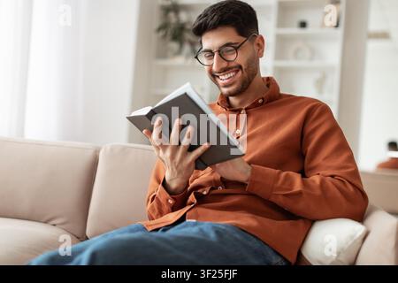 Happy Young Man Reading Book On Couch At Home, Cozy Indoor Setting Stock Photo