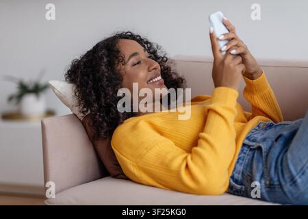 Happy Black Woman Relaxing On Couch While Texting On Smartphone, Living Room Setting Stock Photo