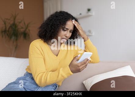 Worried Young Black Woman Using Smartphone At Home, Stressful Situation Stock Photo