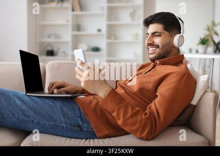 Happy Young Man Using Smartphone And Laptop At Home, Relaxing On Sofa, Business Concept Stock Photo