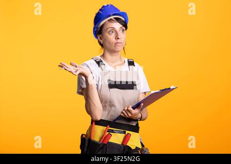 Female builder wearing safety helmet shrugs with uncertain expression while holding clipboard for project notes. Professional contractor stands in tool belt against isolated yellow background. Stock Photo