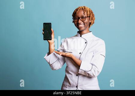 Confident female chef holds mobile phone with green screen against isolated studio backdrop. Cheerful professional cook offers space for culinary ads, restaurant branding and digital food promotions. Stock Photo