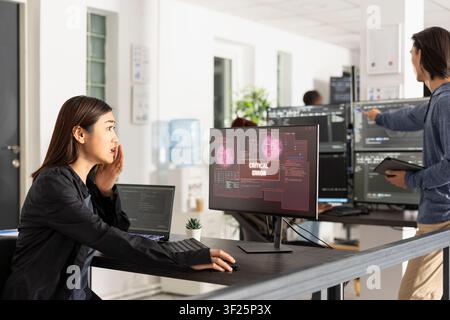 Asian female software engineer reacts in disbelief to security breach alert on desktop monitor, representing hacking, data vulnerability, internet threats and crisis management in modern technology. Stock Photo
