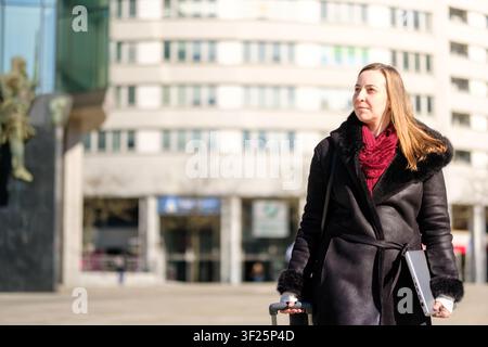 Woman walking urban street with roller luggage, carrying laptop, looking forward Stock Photo