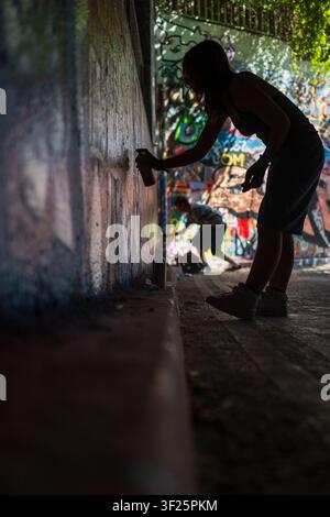 Silhouetted female street artist spray painting mural wall with aerosol can in colorful urban tunnel, London. Stock Photo