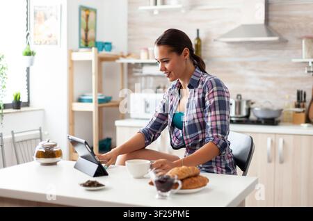 Woman browsing on tablet computer during breakfast in kitchen and holding cup of green tea, working from home using device with internet technology, typing, on gadget during breakfast. Stock Photo