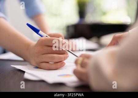 Professional individual with pen in hand, writing on paper during discussion with colleague in office. Close up of hands signing documents on desk as team reviews project and brainstorms ideas. Stock Photo