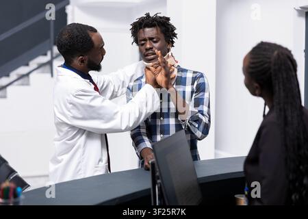 Female receptionist collaborates with doctor and injured man during routine check in at hospital front desk. Physician in lab coat adjusts cervical collar of patient at reception after appointment. Stock Photo