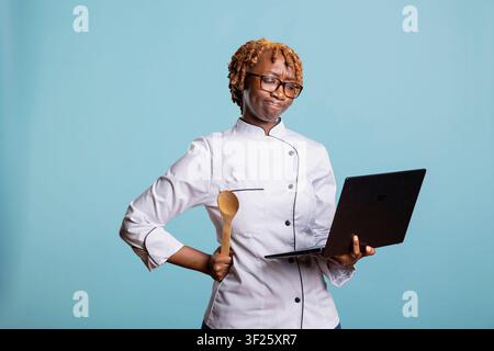Serious black female chef in uniform holds laptop and wooden spoon while planning recipes against blue background. Professional cook represents creativity, learning and modern culinary innovation. Stock Photo