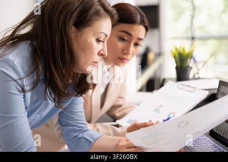Focused female colleagues brainstorm in office, reviewing and preparing project proposals. Asian and caucasian women analyzing charts and financial documents, collaborating on business strategy plan. Stock Photo