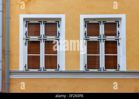 Two symmetrical windows with closed brown wooden shutters and white frames on a textured yellow building facade. Stock Photo