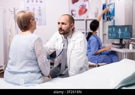 Healthcare specialist using stethoscope in private clinic using stethoscope, Medical practitioner examining lungs of caucasian in modern clinic dressed in white coat. Stock Photo