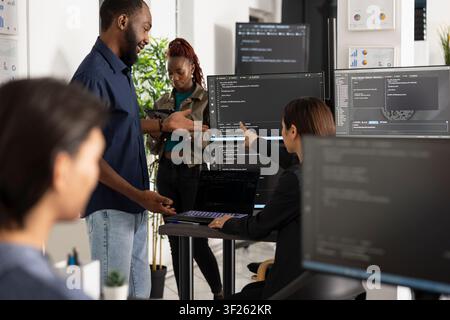 Male and female developers review software data, improving app functionality and company website performance. Diverse coders analyzing HTML scripts on laptop and desktop monitors. Stock Photo