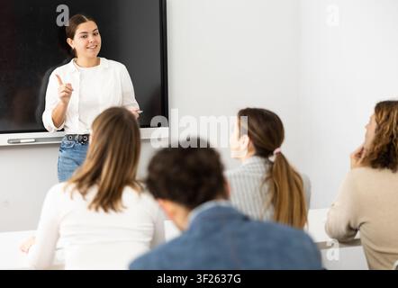 Young female business coach explaining topic while standing against group of people sitting at desks in front of her Stock Photo