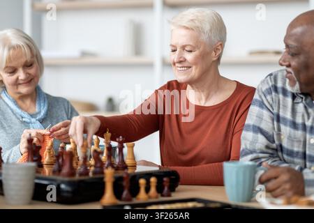 Smiling Senior Women And Man Playing Chess Together In Modern Living Room, Friendly Atmosphere Stock Photo