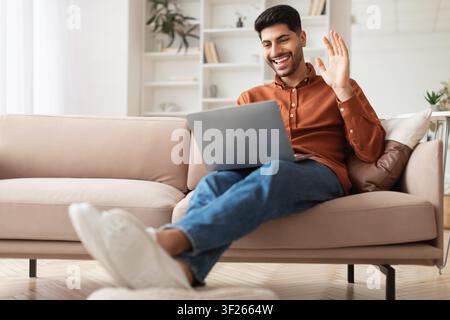 Smiling Young Man Of Middle Eastern Descent Waving At Laptop While Sitting In Cozy Living Room Stock Photo