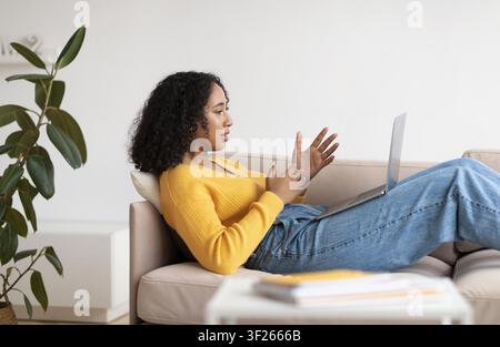 Young Black Woman Engaging In Video Call From Sofa At Home, Remote Work Concept Stock Photo