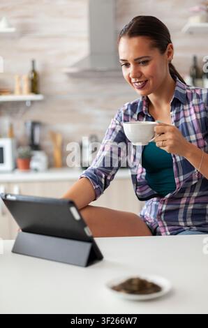 Enjoying cup of green tea while using tablet pc in kitchen. Working from home using device with internet technology, typing, on gadget during breakfast. Stock Photo