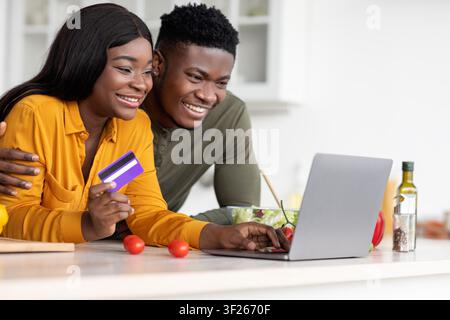 Happy African American Couple Shopping Online At Home, Using Laptop And Payment Card, Kitchen Setting Stock Photo